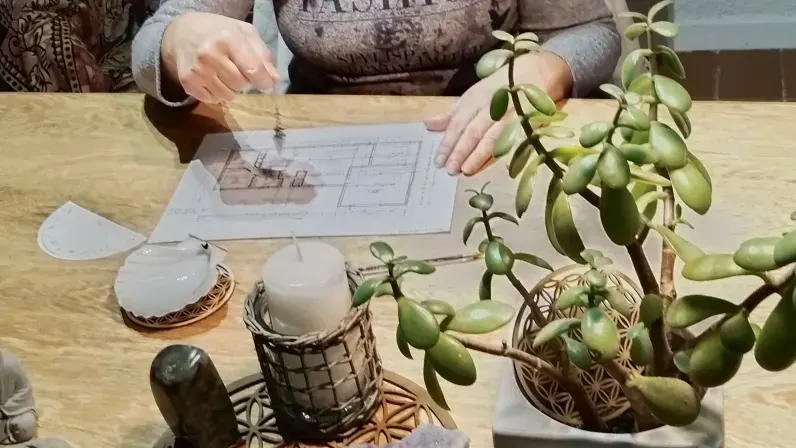 Un pendule tenu au-dessus du plan d'une maison, sur une table avec une bougie et une plante